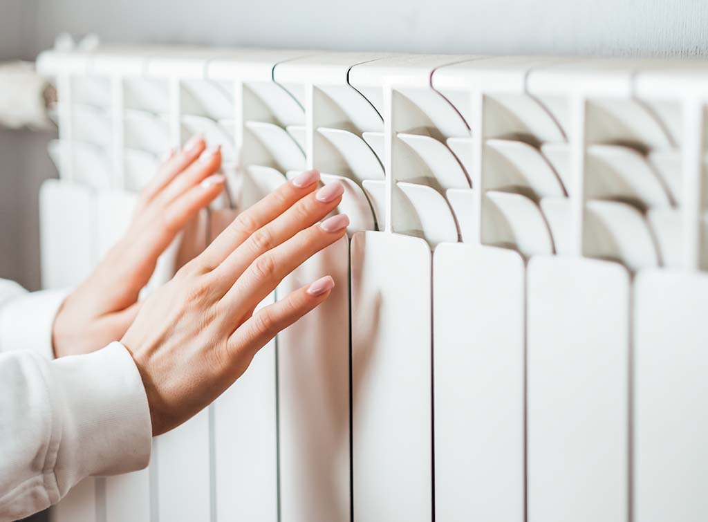 Woman with hands up to radiator for blog: Most economical way to use central heating